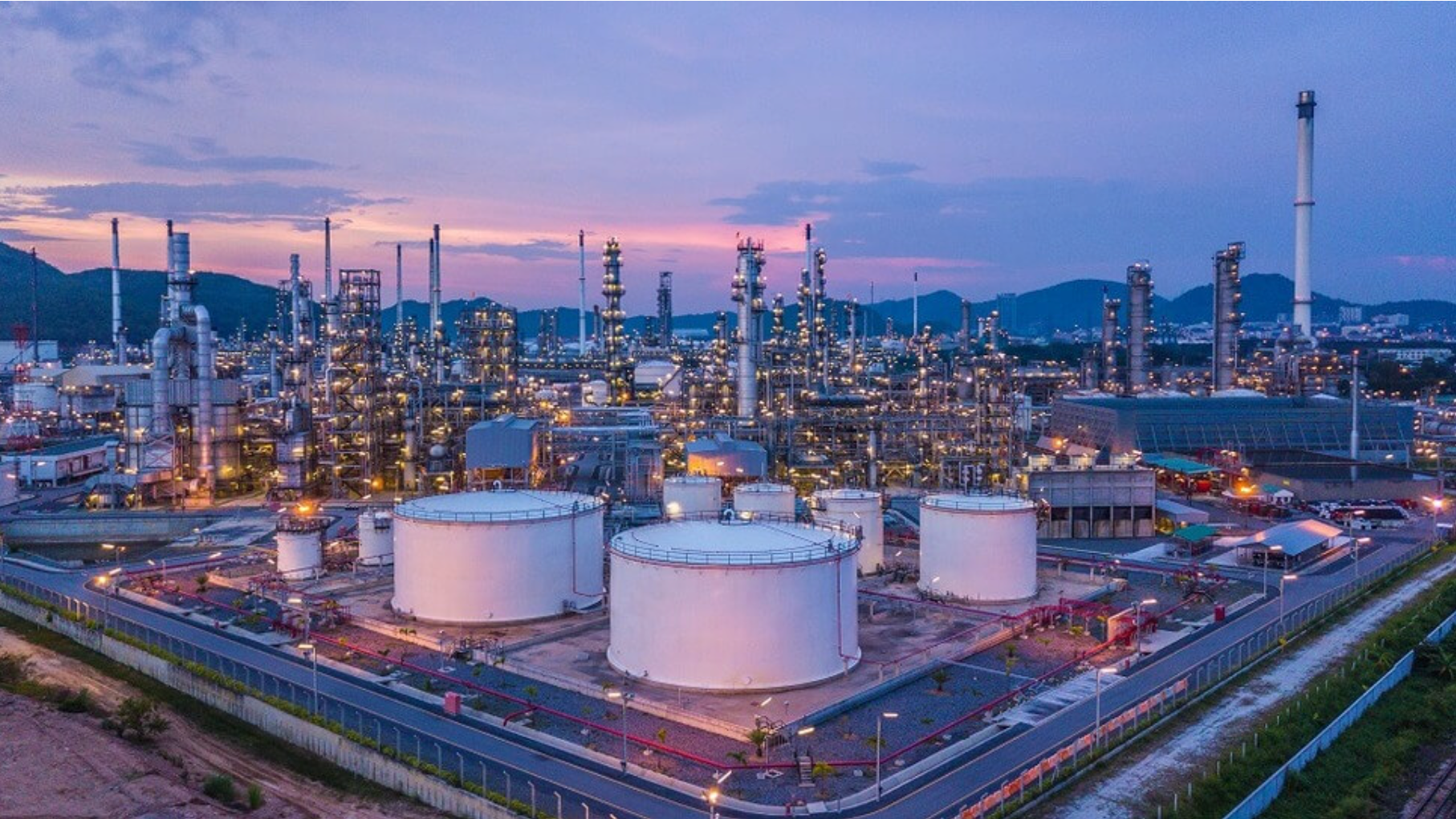 Oil refinery at dusk with large white storage tanks and complex infrastructure.