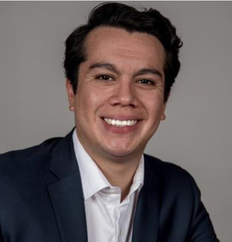 Man in a suit smiles widely, looking directly at the camera against a gray background.
