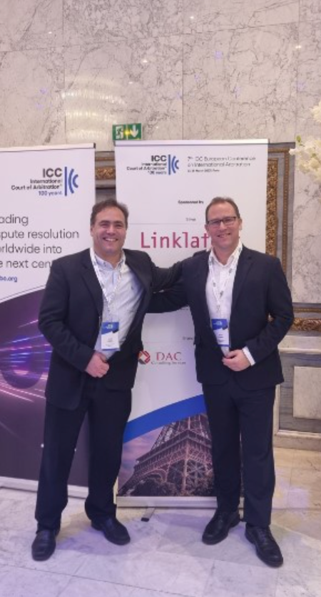 Two men in suits pose at a conference, smiling in front of a banner.