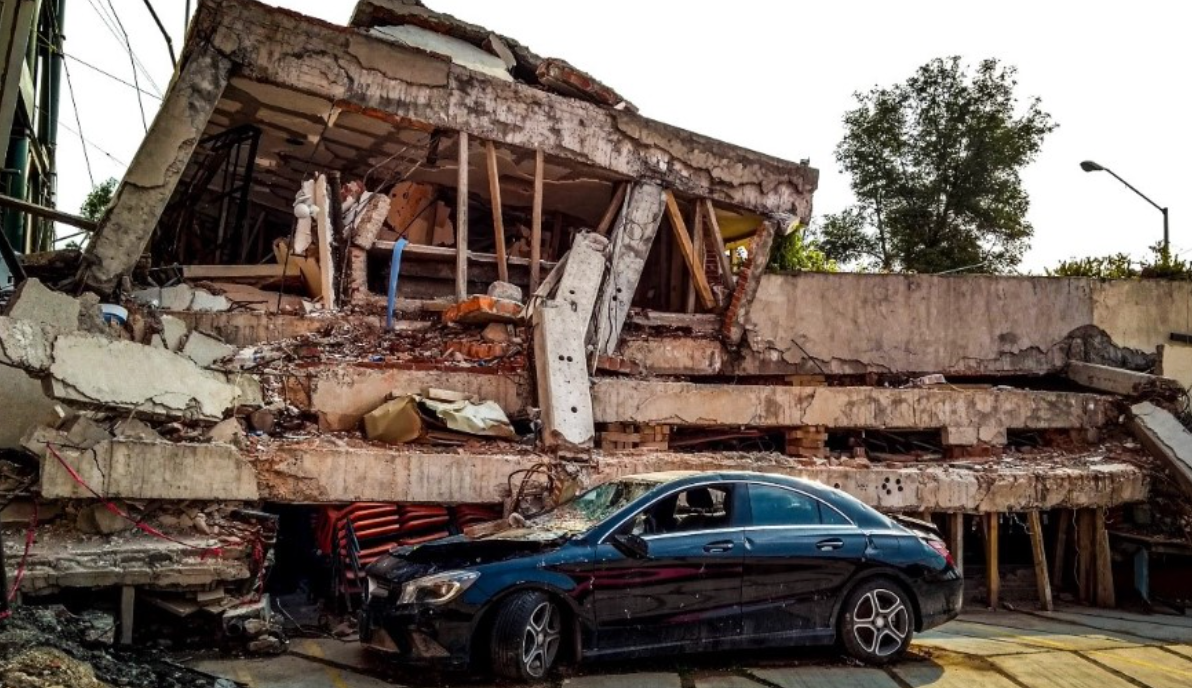 Collapsed building with a black car in front; debris surrounds the structure.