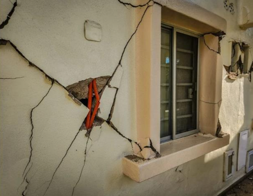 Damaged exterior wall with large cracks and exposed wiring next to a window.
