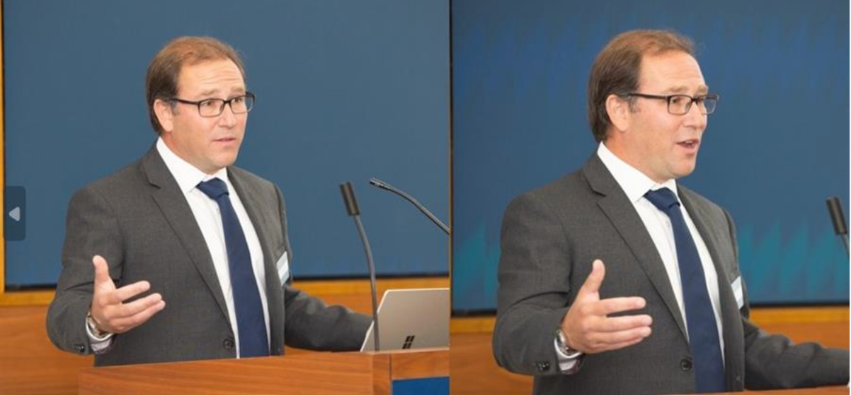 Man in suit giving a presentation at a podium, gesturing with hands.