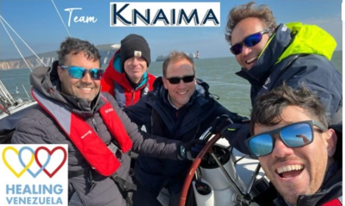 Team KNAIMA on a sailboat with Healing Venezuela logo; five smiling people in outdoor wear.