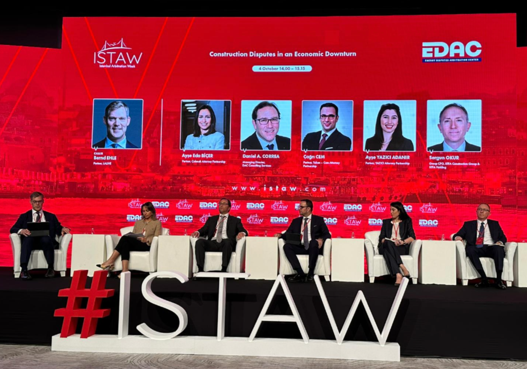 Panel discussion on a red stage with six people in a discussion. Background has speaker headshots.