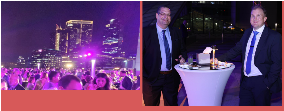 Two men in suits at an outdoor event, people dancing, city lights in background.