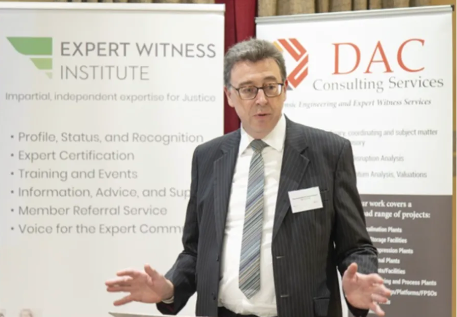 Man speaking at a conference, with banners for Expert Witness Institute and DAC Consulting Services.