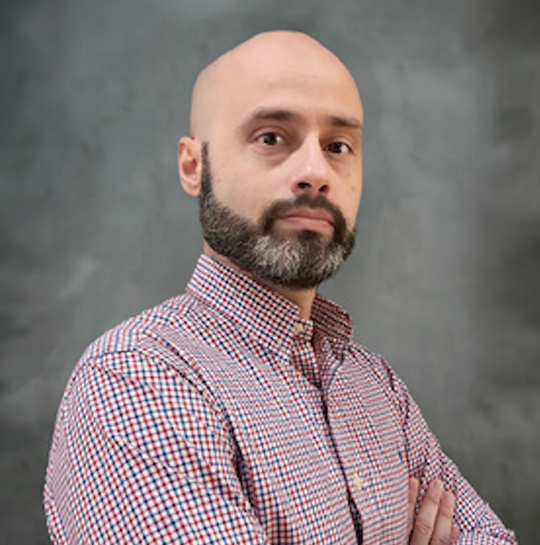 Bald man with a beard, arms crossed, wearing a red and blue plaid shirt, against a gray textured background.