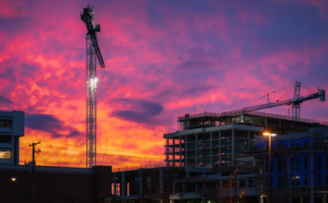 Construction site with cranes silhouetted against a vibrant sunset of pink, orange, and purple hues.