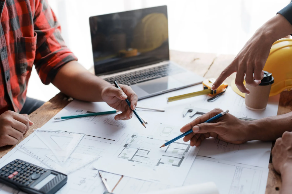 Two people reviewing blueprints, pointing at the designs with pencils. A laptop, calculator, and coffee are on the table.