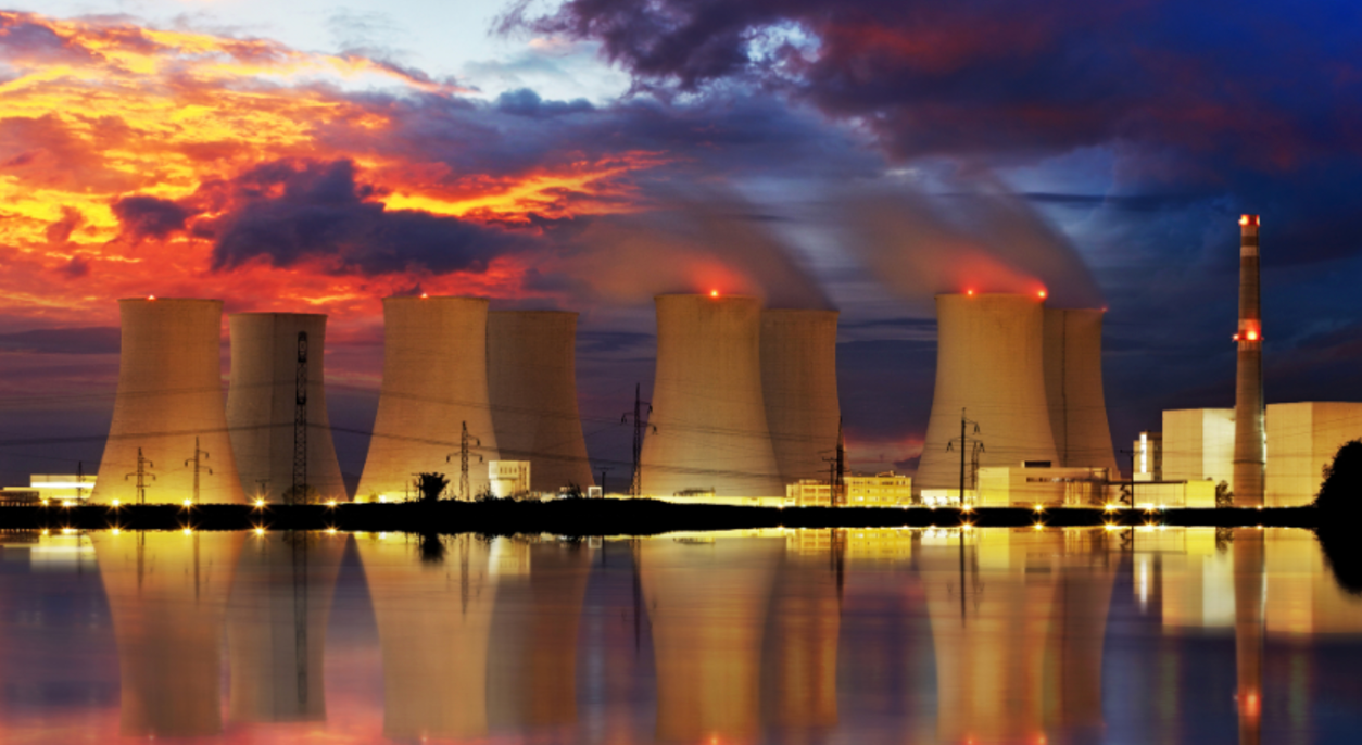 Nuclear power plant at dusk with smoke rising from cooling towers, reflecting in water; colorful sky.