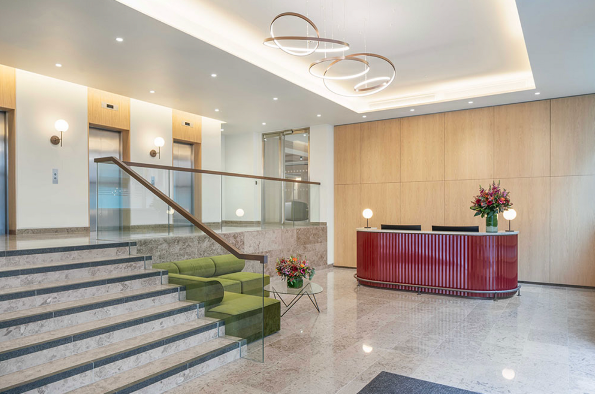 Elegant lobby with marble stairs, green sofa, and a red reception desk. Two elevators and decorative lighting.