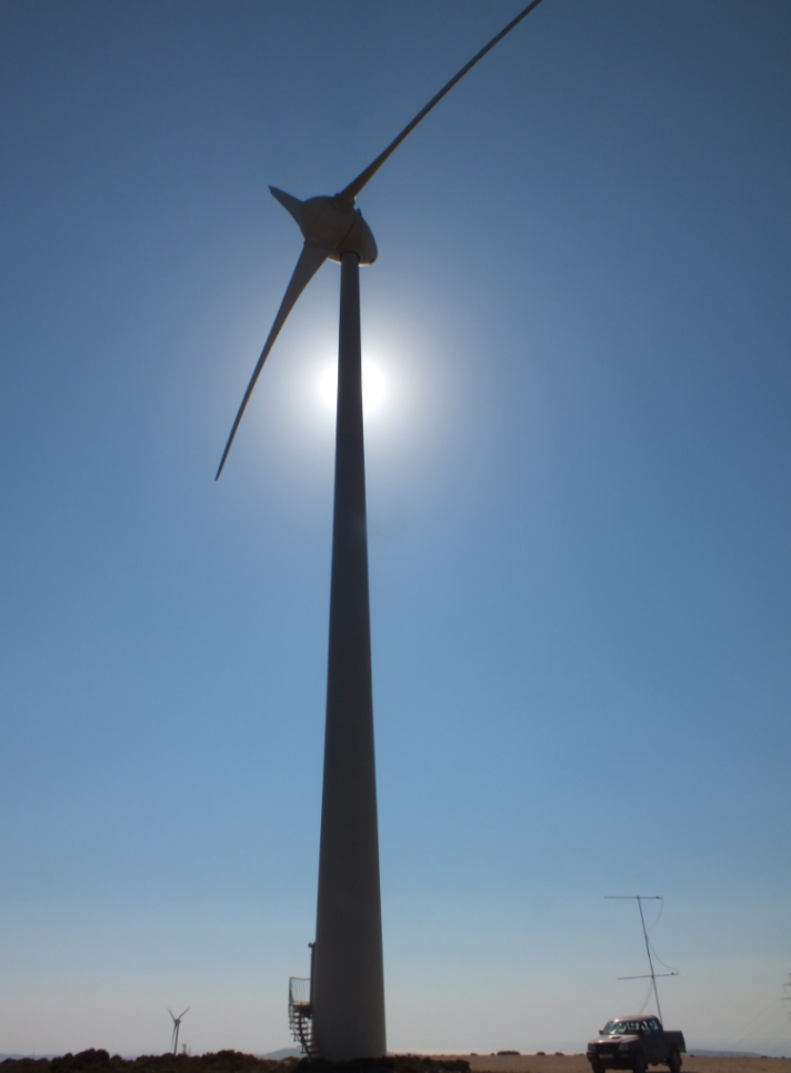 Wind turbine silhouetted against a bright sun, with a vehicle in the foreground under a clear blue sky.