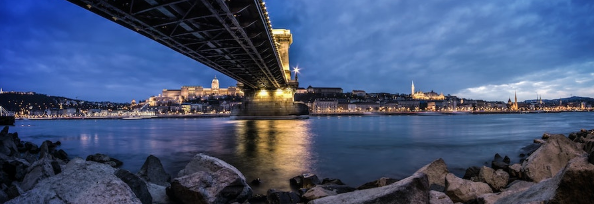 Nighttime view of a bridge over a river, with city lights in the distance and dark, rocky shore in the foreground.