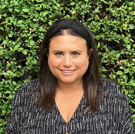 Woman with brown hair and headband smiling in front of a green hedge, wearing a black patterned shirt.
