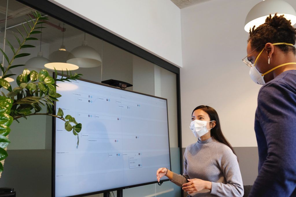 Two people wearing masks discussing a presentation on a whiteboard in an office.