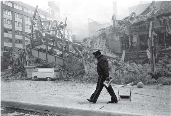 Police officer walks past a collapsed building, debris, and smoke. A van sits nearby.