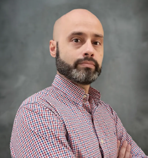 Bald man with a beard, wearing a red and blue plaid shirt, looking at the camera. Against a gray background.