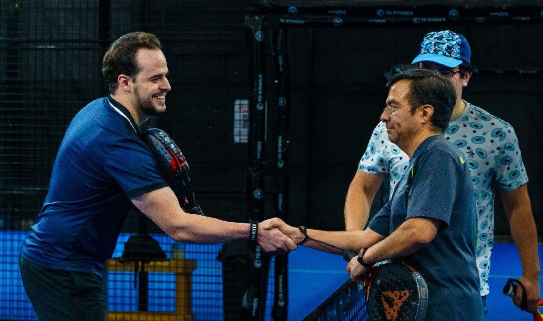 Two men shaking hands on a padel court, a third man standing in the background.