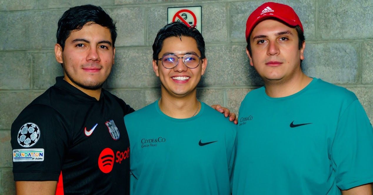 Three men smile, standing close together against a brick wall. Two in teal shirts, one in a black soccer jersey.
