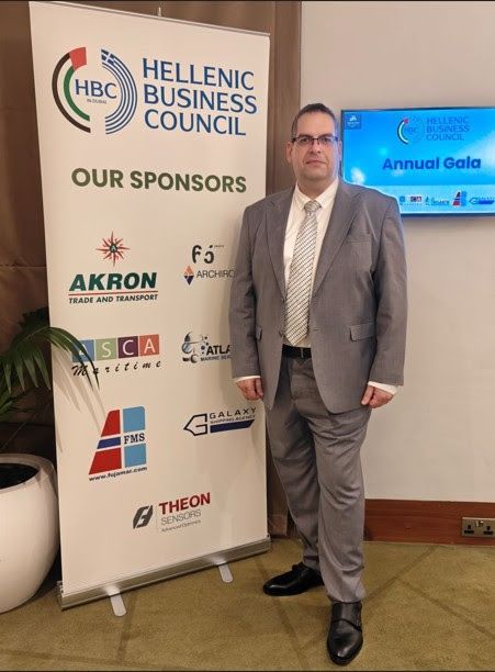 Man in a grey suit stands next to a Hellenic Business Council banner with sponsors.