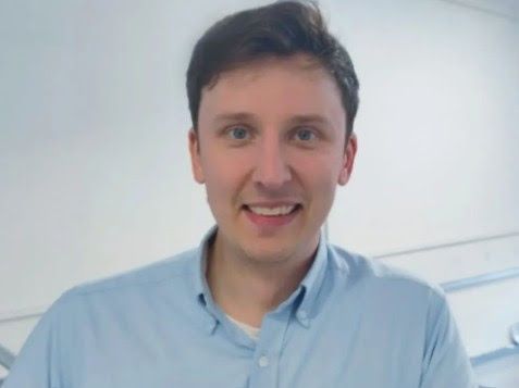Man with dark hair smiling, wearing a light blue button-down shirt, indoors.