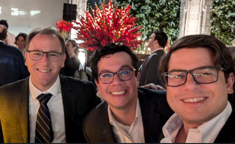 Three men in suits smiling, taking a selfie outdoors with red floral arrangement in background.