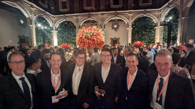Group of people in suits at a formal event, posing in front of a large floral arrangement.