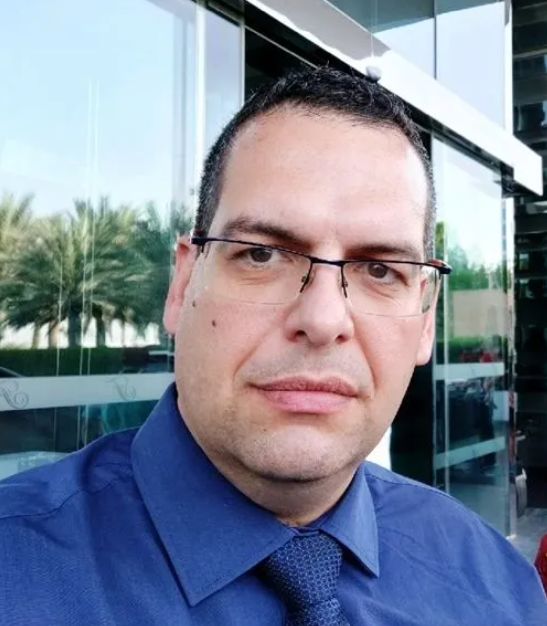 Man with glasses and blue shirt in front of a building with palms.