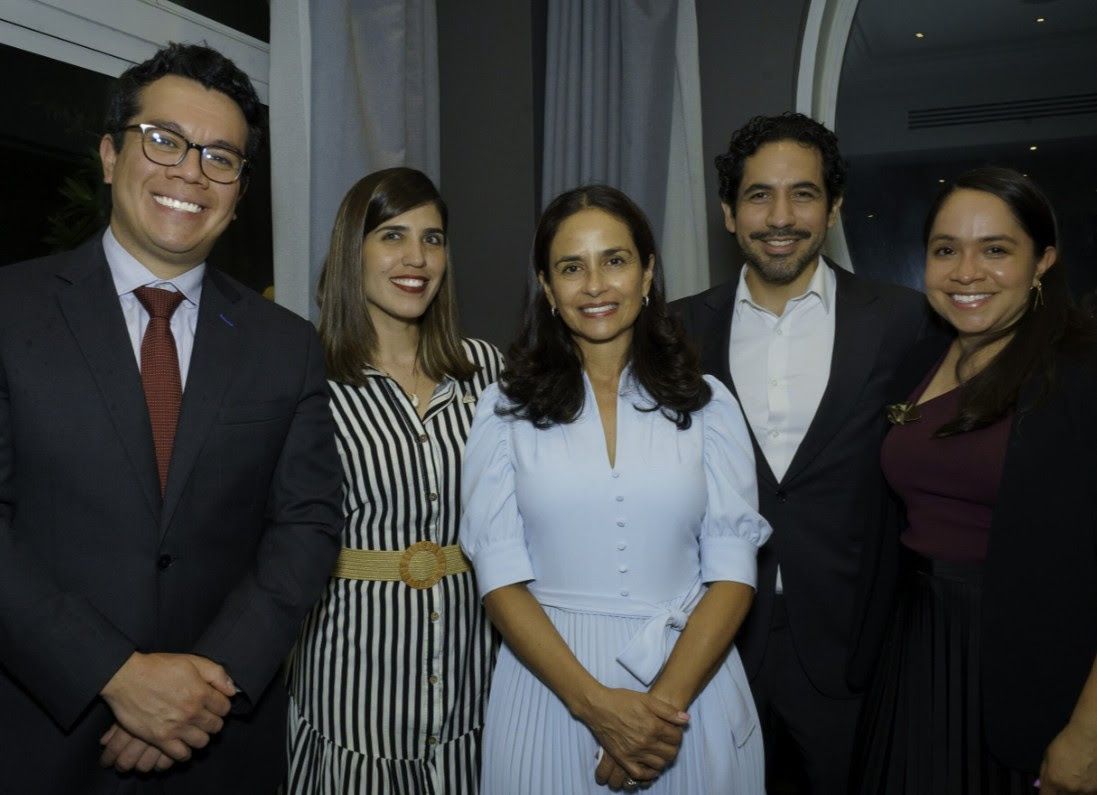 Five people smiling in a room. Men in suits, women in dresses. Neutral setting, indoor.
