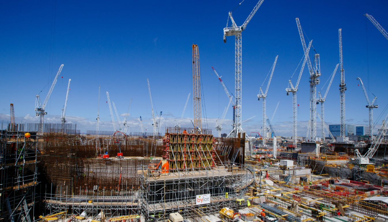 Construction site with numerous cranes against a clear blue sky, showing steel and concrete structures.