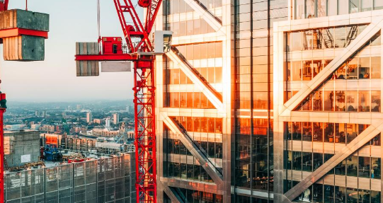 Construction site with red crane lifting materials next to a glass skyscraper, city background.