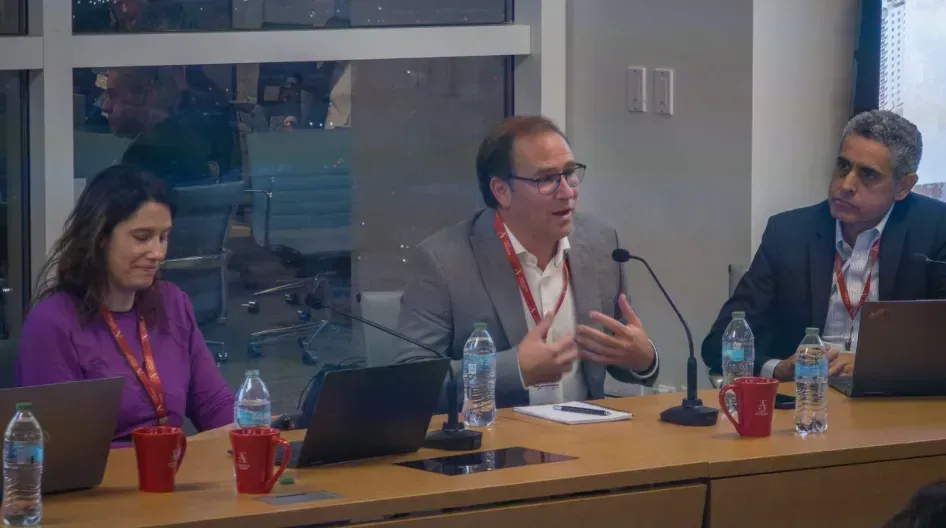 Three people at a table; middle man gestures, speaking into a mic. Two others listen. Indoor setting with a window.