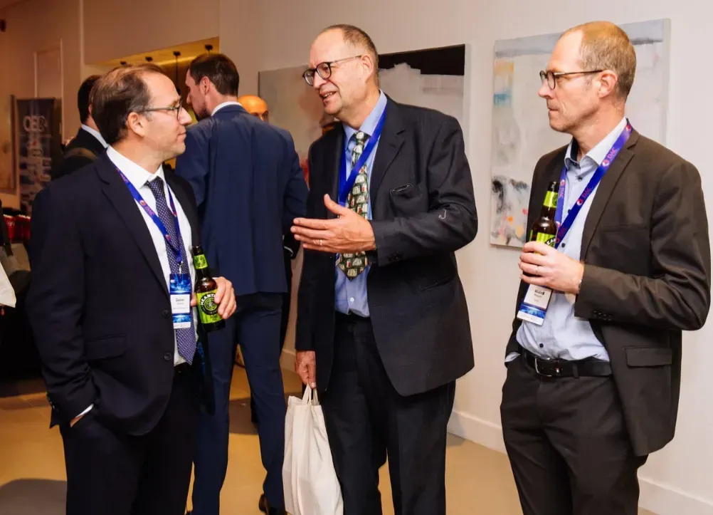 Three men in suits converse at an event, holding drinks and lanyards.
