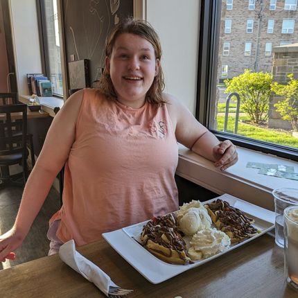 Woman smiling at a table with a dessert plate in front of a window. Light orange top, cafe setting.
