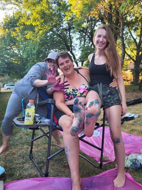 Three women covered in colorful powder in a yard. One seated person shows off blue feet, while the others smile.