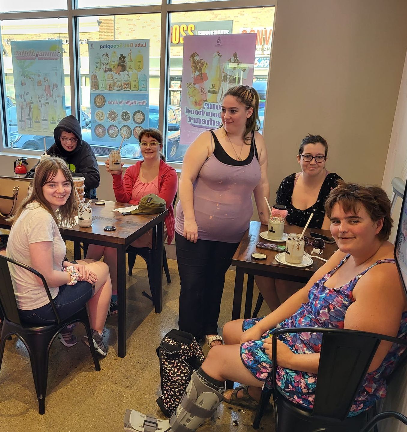 Six women at tables in a cafe. Some are smiling, holding cups. Posters on wall.