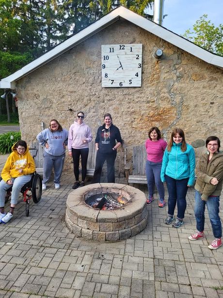 Group of women standing around a fire pit on a patio.  Stone building with a clock in the background.