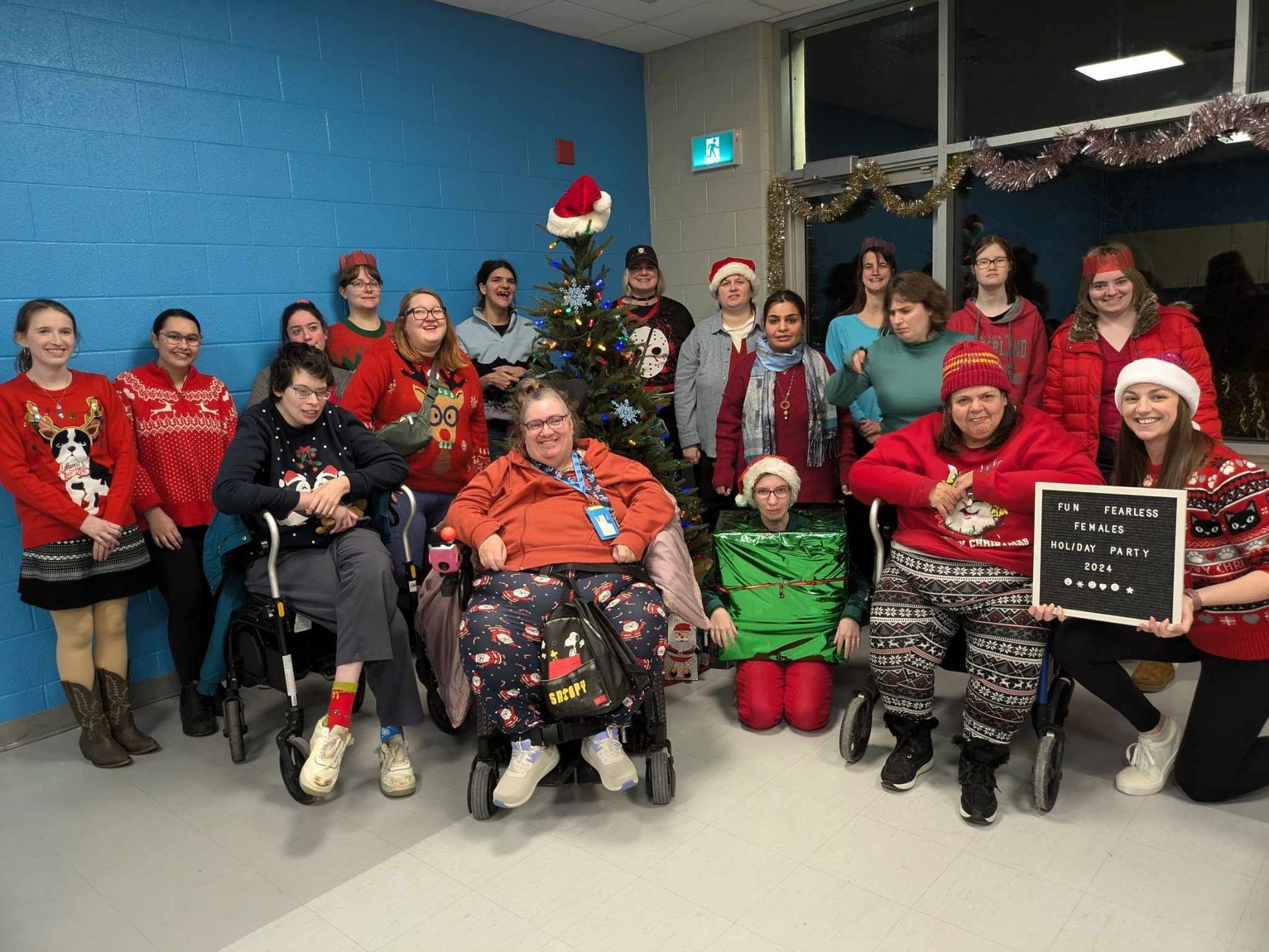 Large group of women in festive outfits around a Christmas tree, some using wheelchairs, in front of a blue wall.