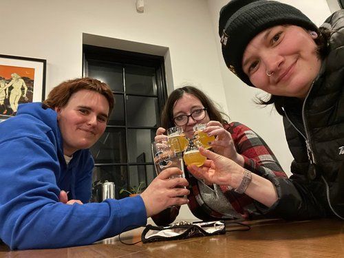 Three women indoors, toasting drinks. One in a blue hoodie, one in a plaid shirt, and one in a beanie.
