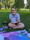 Woman sitting cross-legged in a park, holding a colorful painting; art supplies and painted canvas on ground.