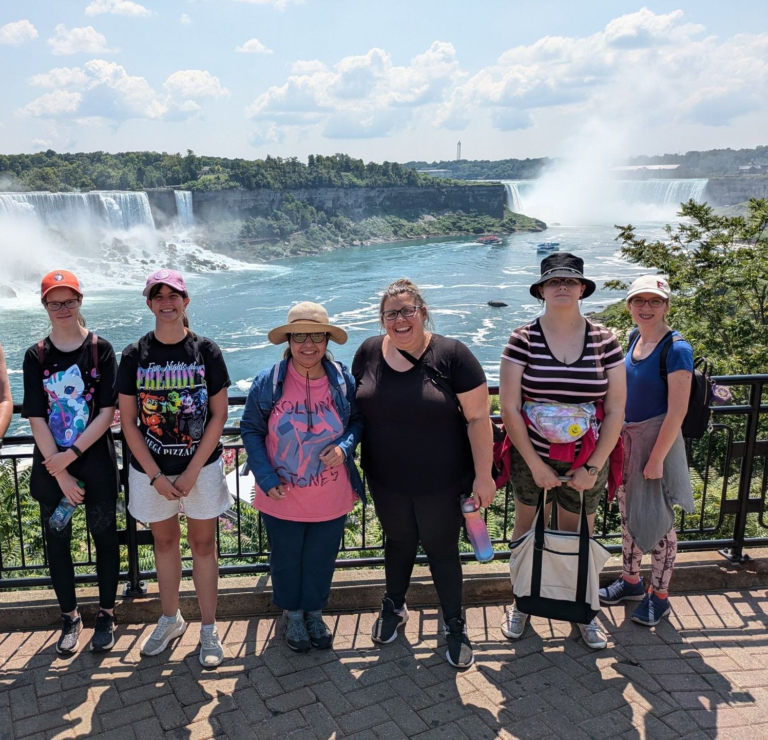Six women pose in front of Niagara Falls. Waterfalls in the background, sunny day.
