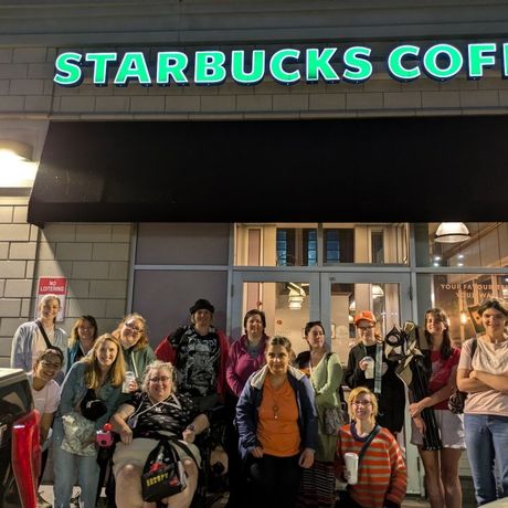 Group of womenin front of a Starbucks, some holding drinks. Store sign lit up. Nighttime.