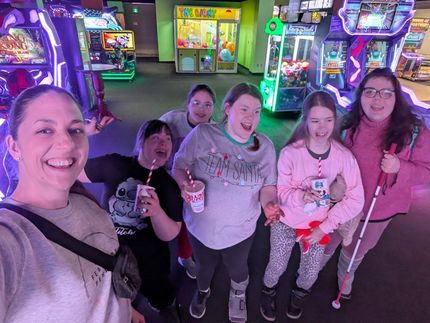 Six girls smile at an arcade, holding drinks. One uses a white cane. Purple and green lighting.