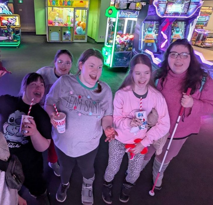 Group of girls at an arcade: laughing, holding drinks, one with a cane. Neon lights and game machines in background.