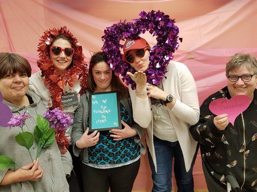 Group of five women posing with Valentine's Day props, including heart-shaped wreaths and holding flowers.
