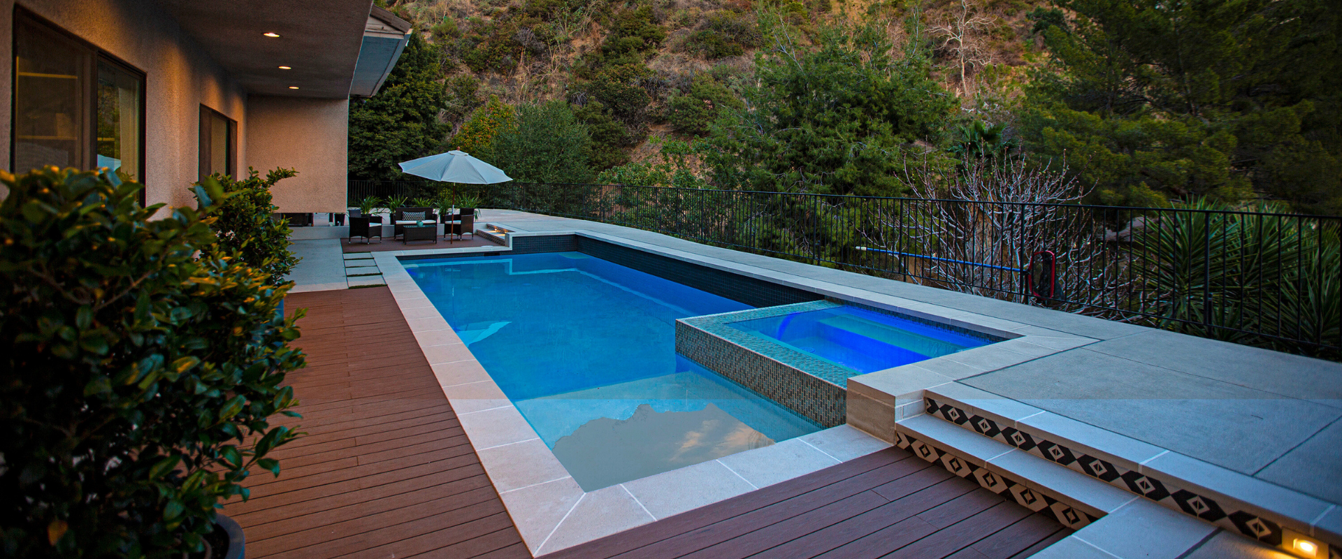 A rectangular swimming pool and hot tub surrounded by a wooden deck, set against a backdrop of greenery.