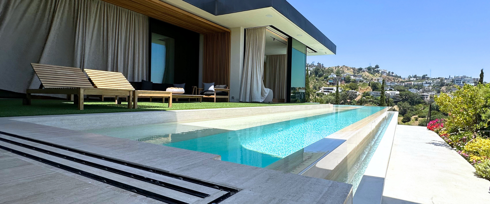 An infinity pool with a view of a hillside. Lounge chairs on a patio with a building on the left.