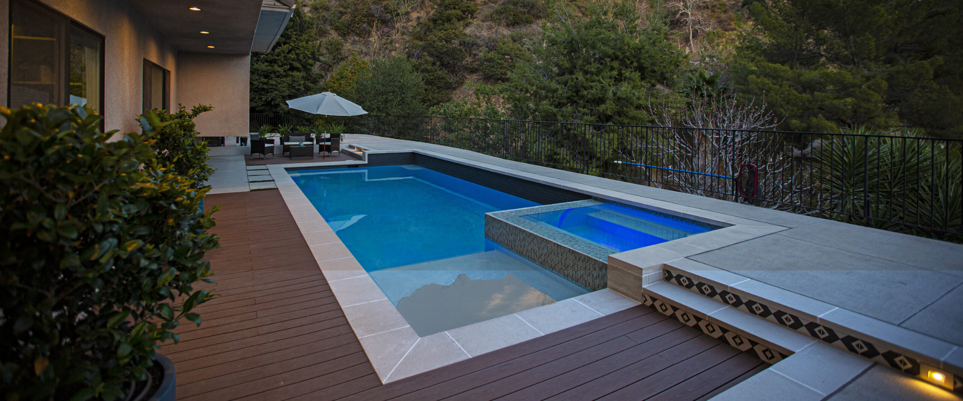 Modern backyard pool with blue water, wooden deck, hot tub, and trees at dusk