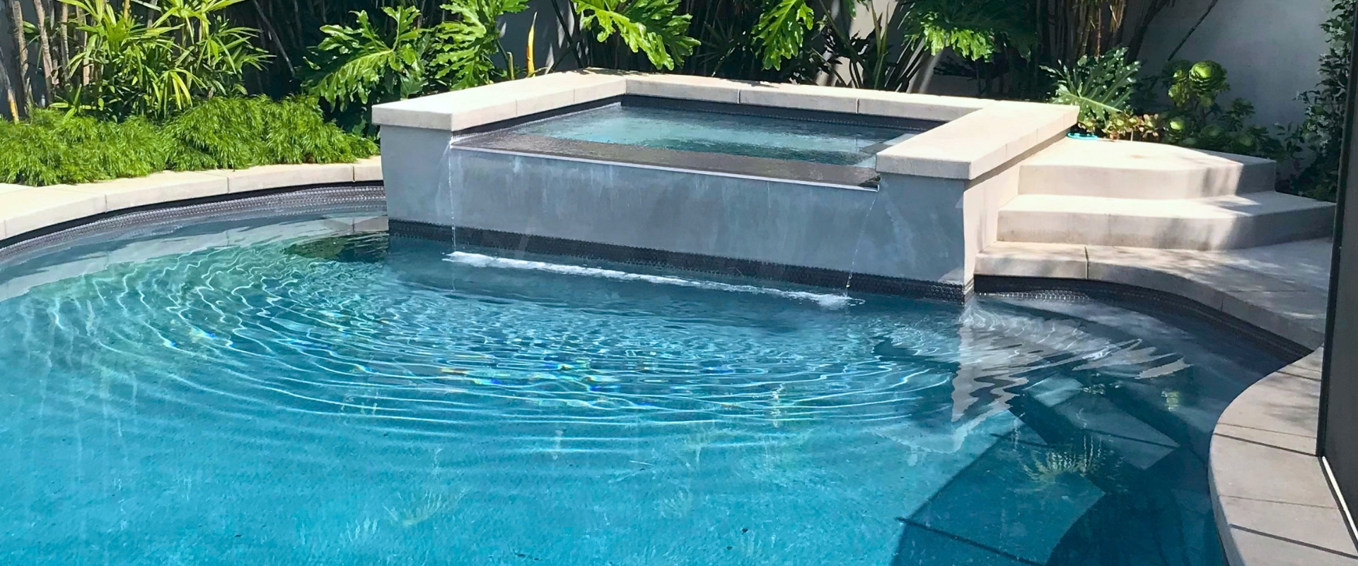 Backyard pool with blue water and a raised stone spa surrounded by greenery