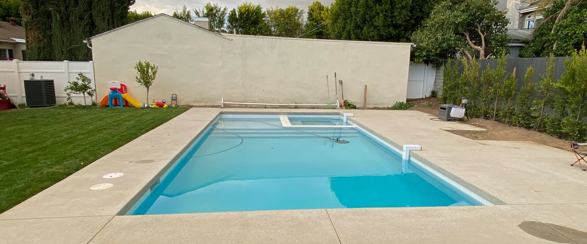 Backyard swimming pool with blue water, beige deck, and green lawn beside a tall wall